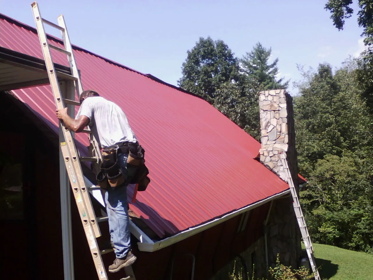 Roofer on a ladder beside a red metal roof and stone chimney — Soffit & Fascia Repair by Roofing Brothers in Mosinee